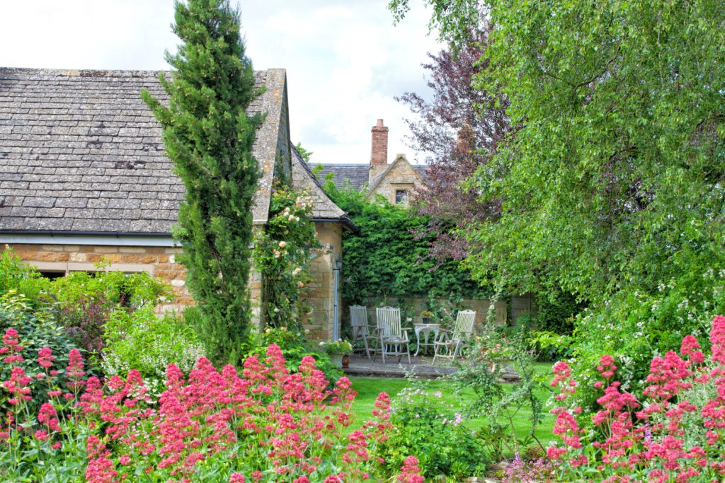 Enclosed patio with chairs and table in charming Cotswolds cottage garden full of flowers in bloom, mature trees .