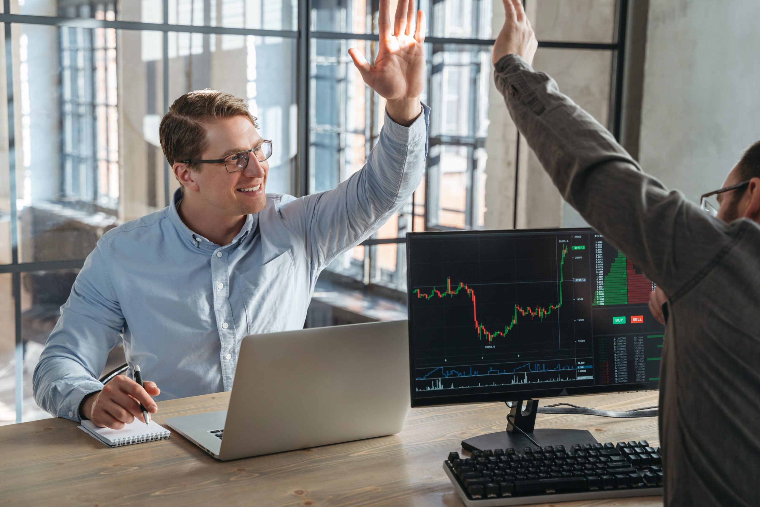Two partners trade investors giving high five to each other after fulfilling good deal, buying crypto currency at stock market at low price, sitting opposite each other in front of computers